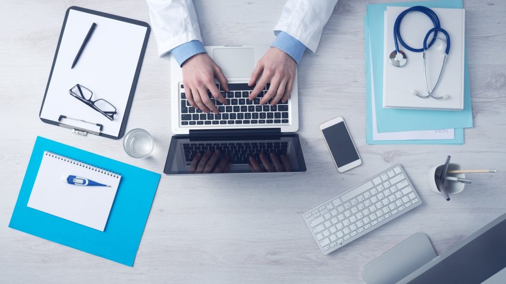 A pair of hands typing on a laptop surrounded by a pens and pencils, cellphone, notebook, and stethoscope.